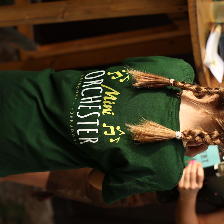 A girl with braided hair wearing a green shirt with the text 'Kanddorf, Ebersdorf' and musical notes on it.