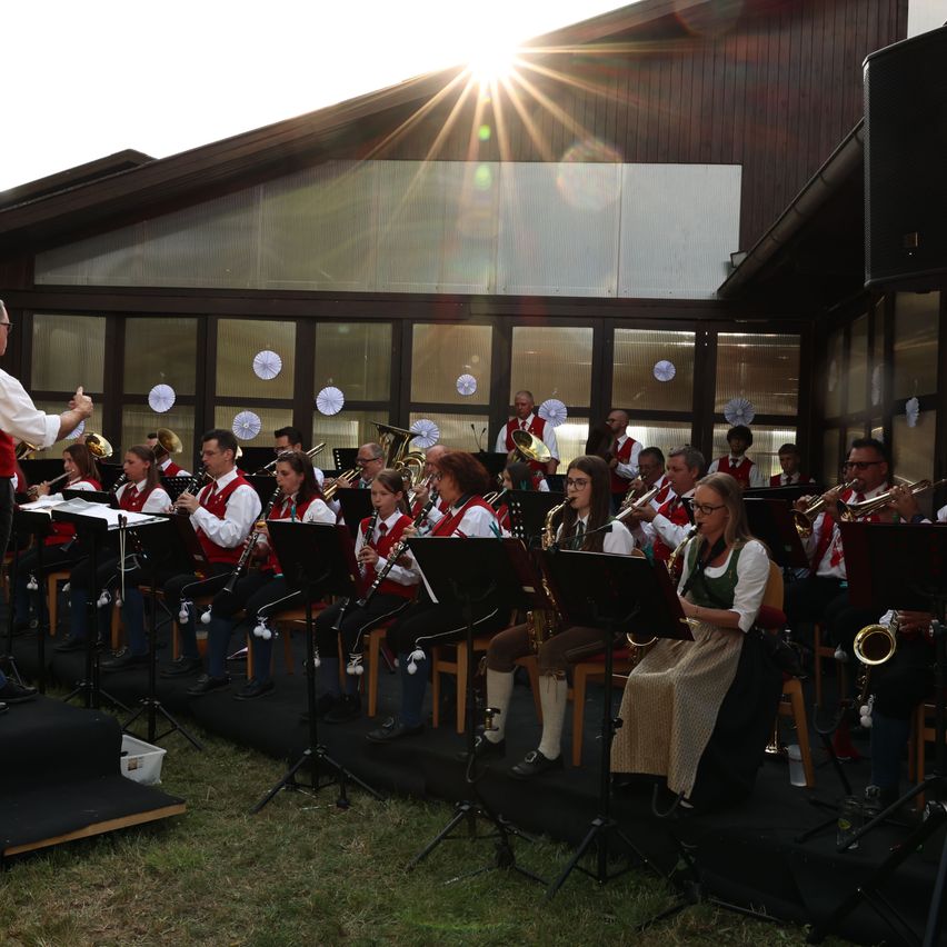 A brass band performs outdoors in front of a modern building with large glass windows. The musicians are seated and standing, playing various instruments. The sun is shining brightly behind them.