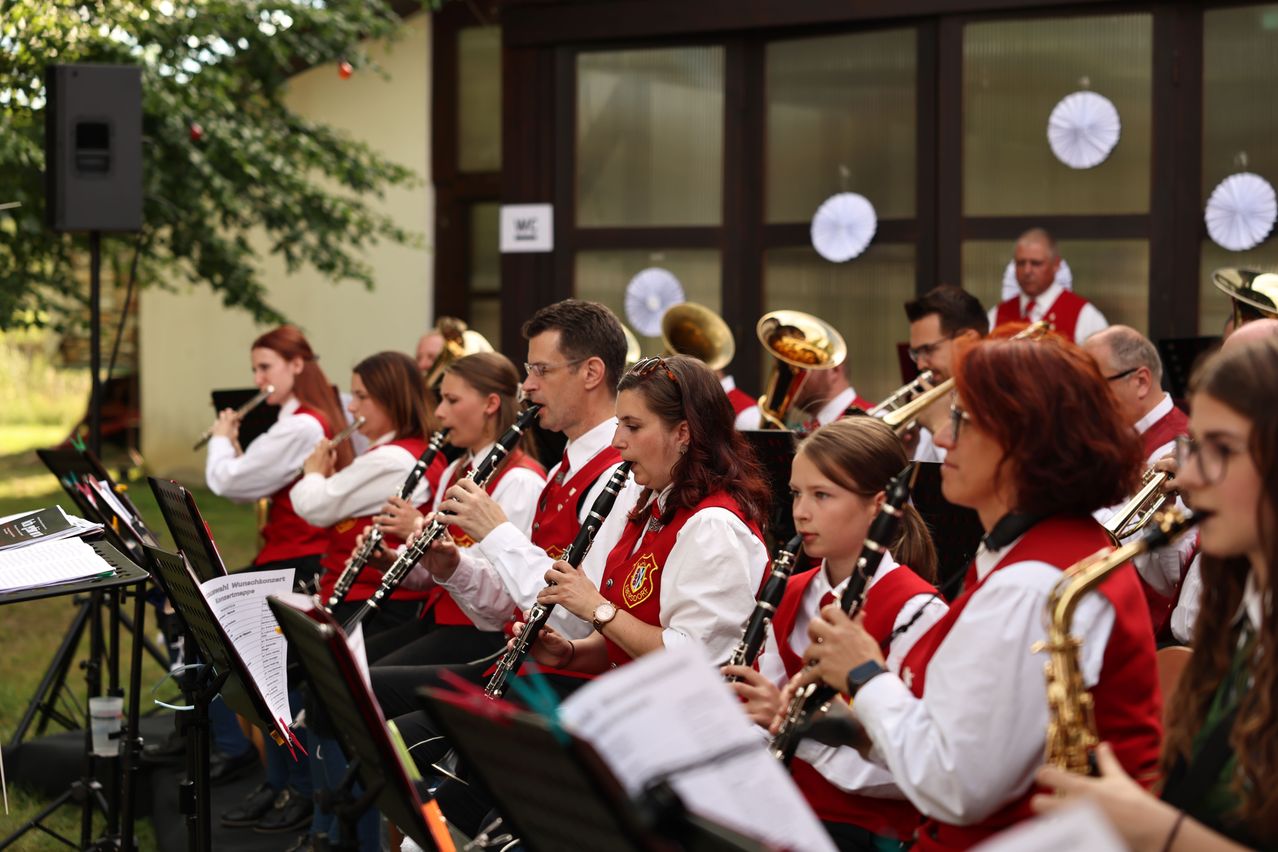 A group of musicians in red vests are playing various instruments, including clarinets and trumpets, in an outdoor setting.