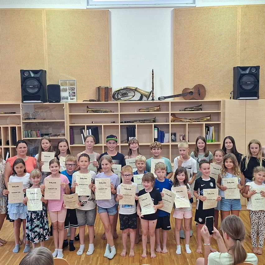Eine Gruppe von Kindern und Erwachsenen steht in einem Raum und hält Urkunden in der Hand, mit Musikinstrumenten in den Regalen im Hintergrund.