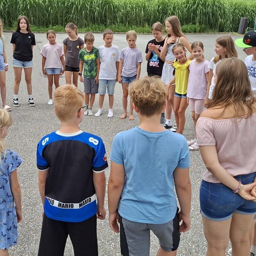 Eine Gruppe von Kindern und Teenagern steht in einem Kreis, wahrscheinlich für ein Gruppenfoto, in einem Außenbereich mit hohem Gras im Hintergrund.