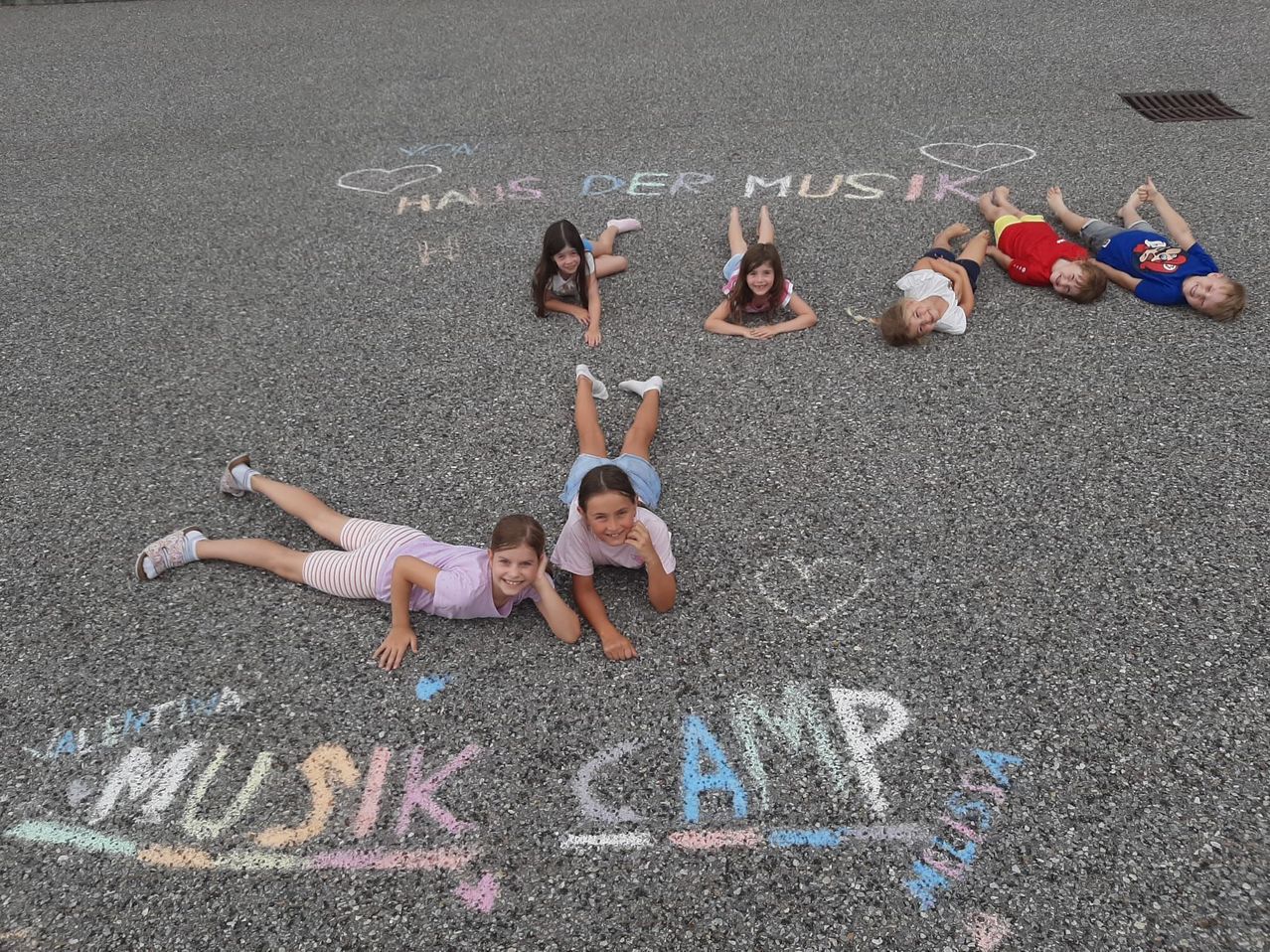 Fünf Kinder liegen auf dem Boden und bilden eine Herzform, mit Kreideschrift um sie herum, die 'Music Camp' und 'Habt ihr der Musik' lautet. Sie lächeln und scheinen für ein Foto zu posieren.