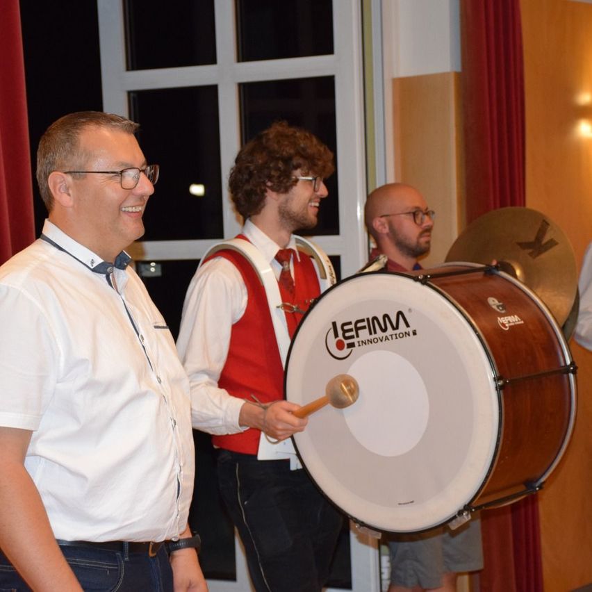 Three men in a room with a drum, one smiling, another holding a drumstick, and the third standing behind the drum.