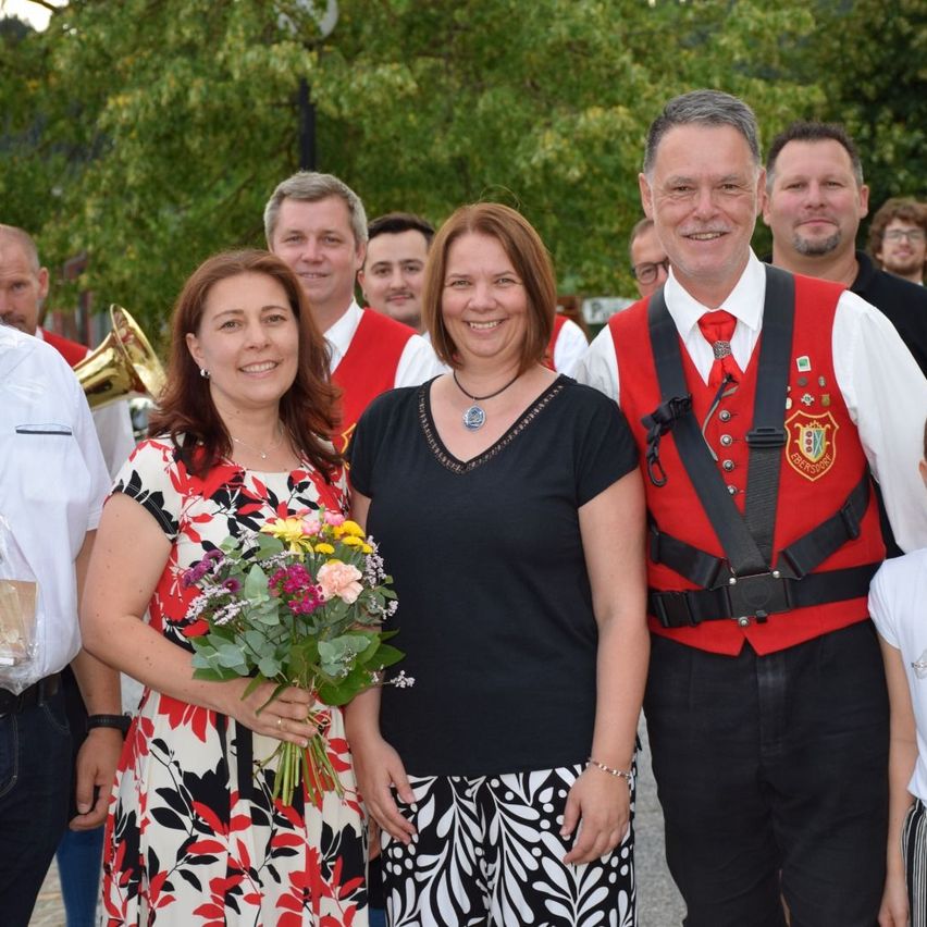 A group of people dressed in traditional attire, smiling and posing for a photograph. Two women hold flowers, and a man wears a red vest with a logo.