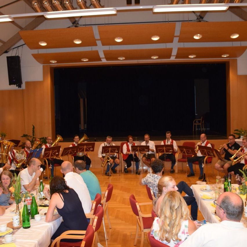 A group of musicians in red vests are performing on stage in a hall with an audience seated in front of them. The musicians are playing various instruments, and the audience is enjoying the performance.