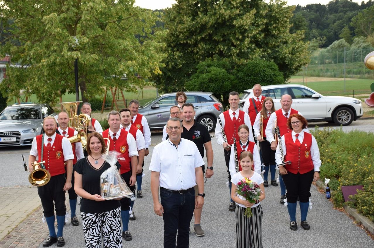 A group of musicians in red vests and white shirts, some holding instruments, stand in a line with a woman holding a gift basket and a girl with flowers in front of them.