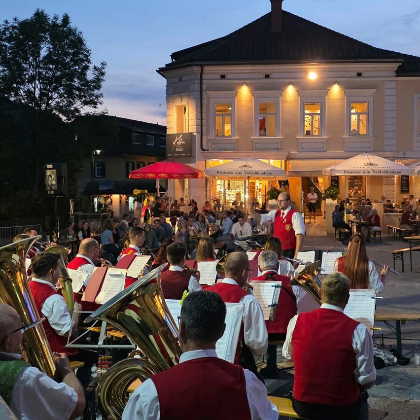 A band in red vests plays instruments in an outdoor setting at dusk, with a crowd of people in the background and a building with lit windows.