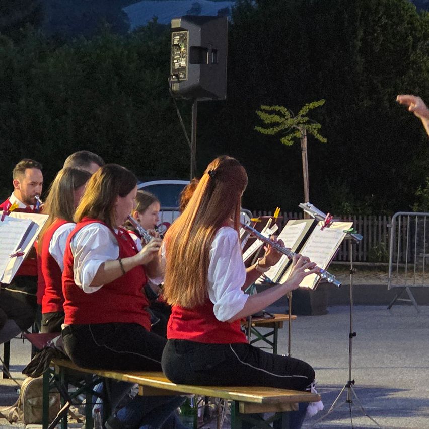 A group of musicians in red vests and black pants are playing flutes on a bench in an outdoor setting. A speaker and a small tree are behind them.