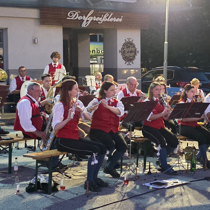A brass band performs outdoors in front of a building with a sign reading 'Dorfreig Sterei'. The musicians are seated on benches, playing various instruments including trumpets, trombones, and saxophones.