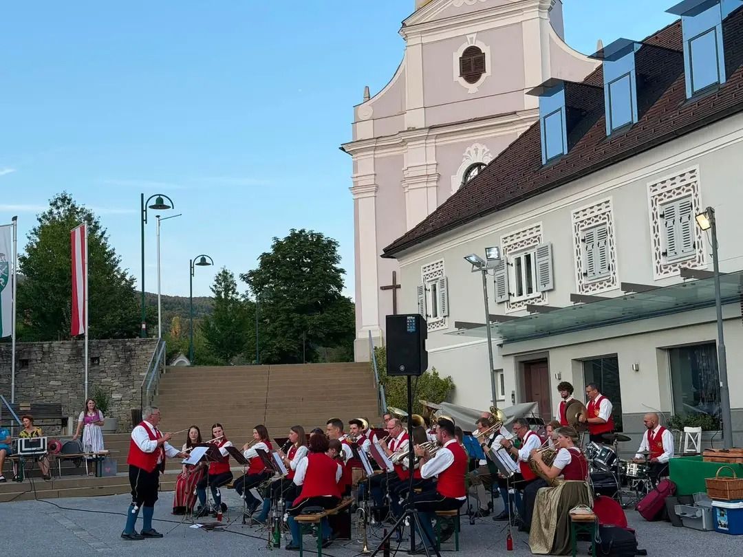A brass band performs outdoors in front of a church, with musicians in traditional attire. The setting includes a large building with ornate windows and a clear sky.