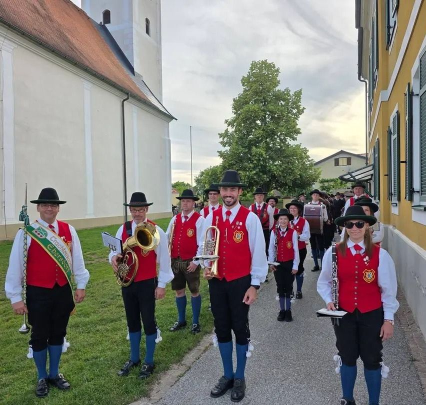A group of musicians dressed in red vests and black hats walk down a street, holding brass instruments and smiling for the camera.