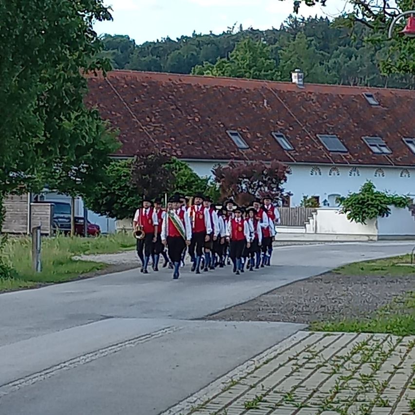 A group of people in matching uniforms are marching down the road in front of a house with a red roof.