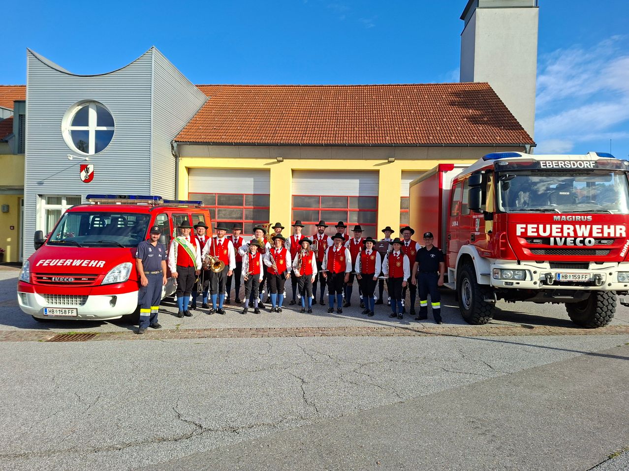 A group of firefighters in red uniforms and hats pose for a photo in front of a fire station with red fire trucks on a sunny day.