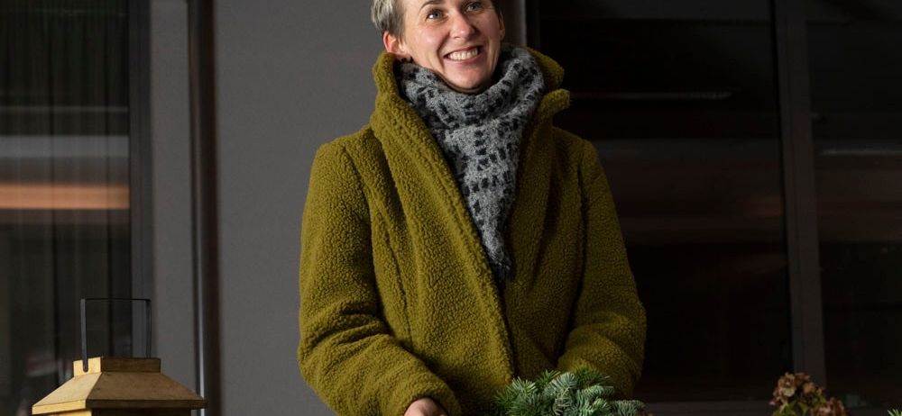 Eine Frau mit kurzen Haaren lächelt und hält einen Weihnachtskranz vor einem Gebäude. Sie trägt einen grünen Mantel und einen Schal.
