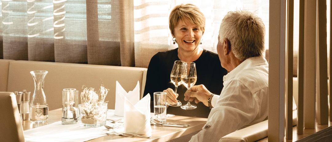 Ein Paar sitzt an einem Tisch, lächelt und hält Weinglas in der Hand. Sie sind formell gekleidet. Der Tisch hat eine weiße Tischdecke, eine Blumenvase und Gläser. Hinter ihnen befinden sich Fenster mit Vorhängen.