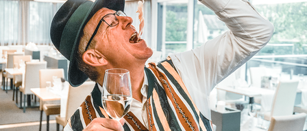 Ein älterer Mann in Hut und Brille isst ein Stück Essen mit offenem Mund, während er ein Weinglas in einem Restaurant hält.