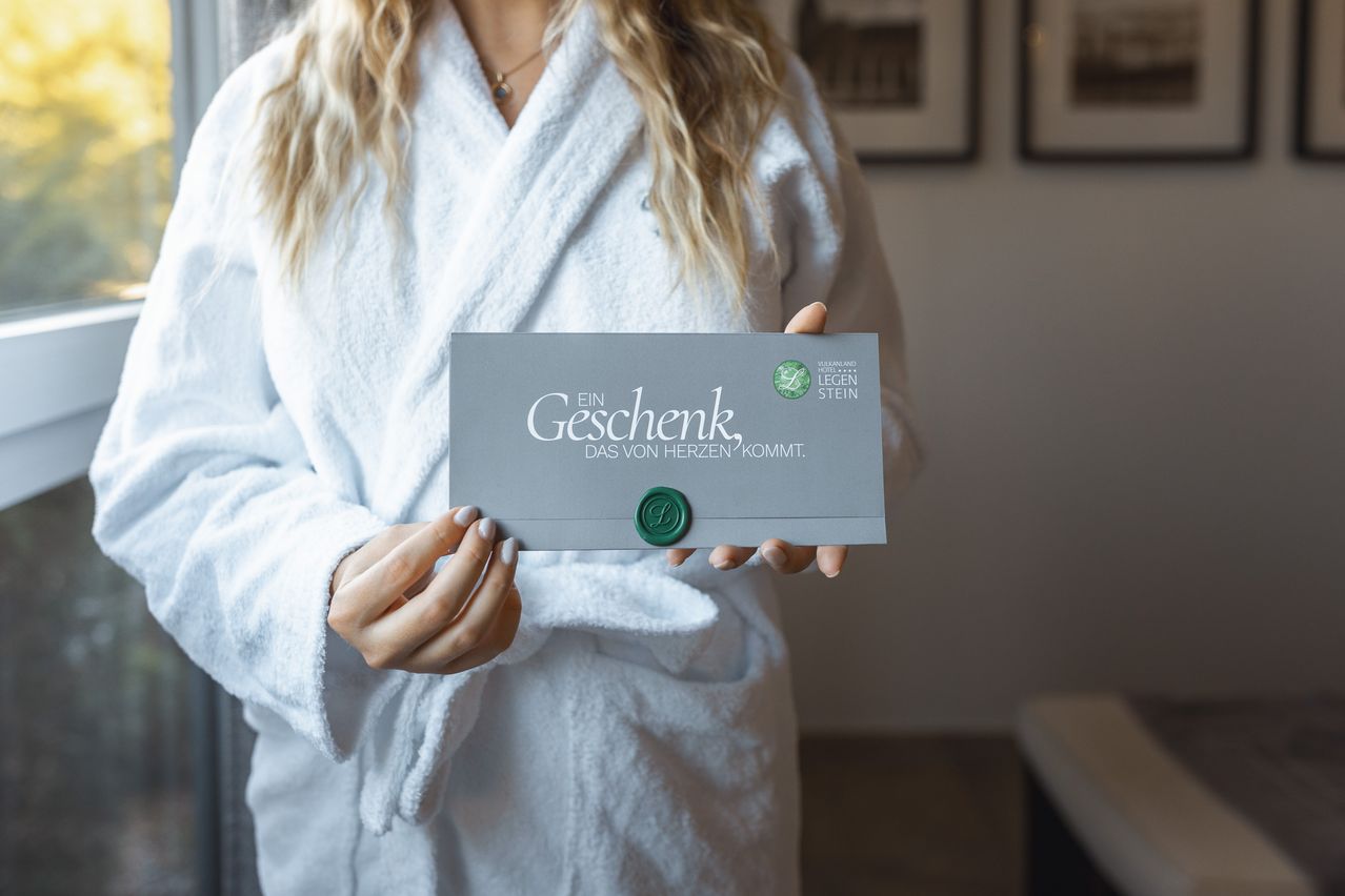 Eine Frau in einem weißen Bademantel hält einen grauen Umschlag mit einem grünen Wachssiegel. Der Umschlag trägt 'Ein Geschenk' in fetten Buchstaben. Das Bild befindet sich in einem Raum mit weißen Wänden.