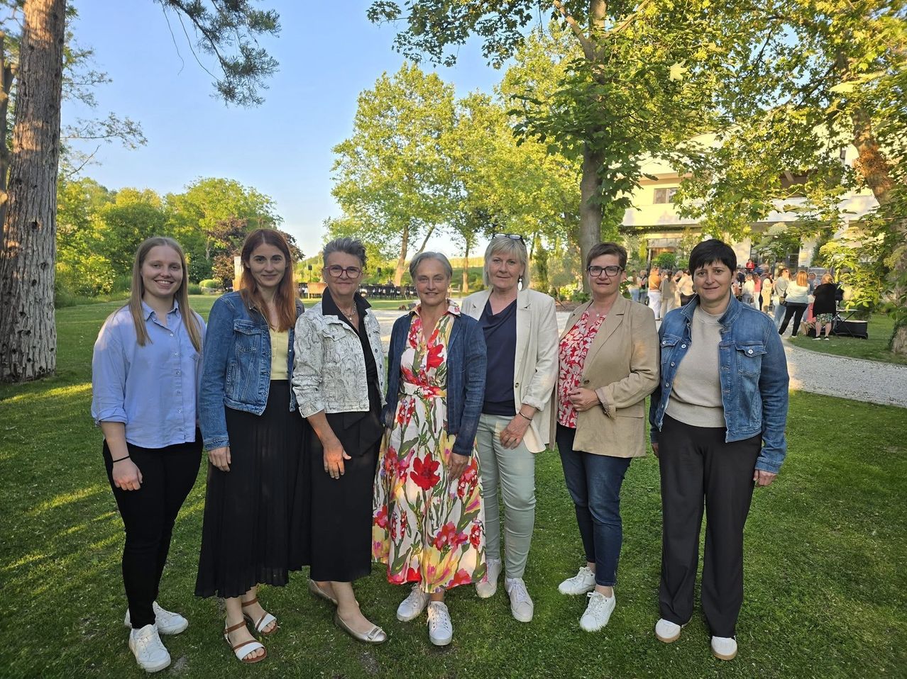 Sechs Frauen stehen auf einer Rasenfläche und lächeln für ein Foto. Dahinter sind Bäume und ein Gebäude mit Menschen in der Ferne.