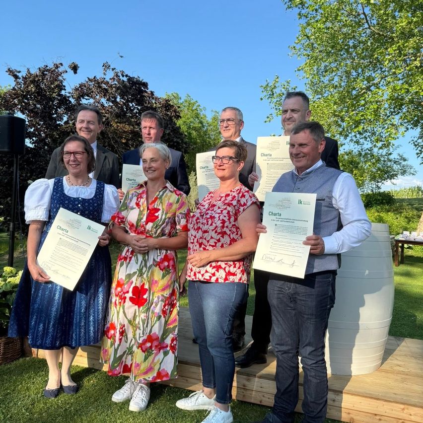 Sieben Erwachsene posieren mit Zertifikaten in einem Garten. Drei Frauen und vier Männer stehen auf einer Holzplattform. Im Hintergrund befinden sich grüne Bäume.