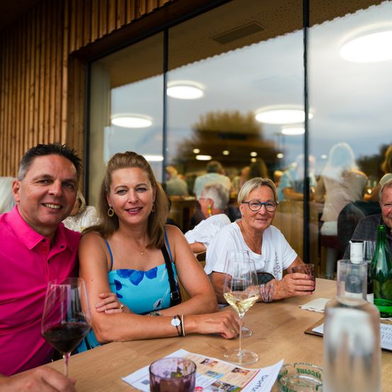 Eine Gruppe von Menschen, darunter ein Mann und zwei Frauen, sitzt an einem Tisch und lächelt. Auf dem Tisch stehen Weingläser und Flaschen. Sie befinden sich in einem Restaurant mit Glaswänden und Holzakzenten.