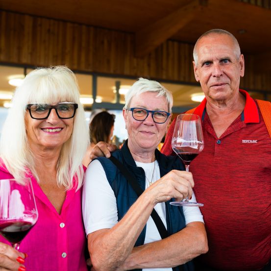 Drei Personen stehen zusammen in einem Raum und halten Weingläser. Ein Mann und eine Frau lächeln, während die Frau eine Brille trägt und ein rosa Hemd trägt.
