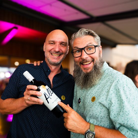 Zwei Männer lächeln und lachen, einer hält eine Weinflasche mit der Aufschrift 'All', während der andere darauf zeigt. Sie sind drinnen, vermutlich bei einer Veranstaltung.