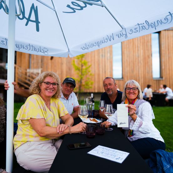 Vier Erwachsene sitzen lächelnd und mit Weingläsern an einem Tisch im Freien. Sie sind unter einem großen weißen Sonnenschirm mit blauer Schrift. Hinter ihnen ist ein hölzernes Gebäude zu sehen.