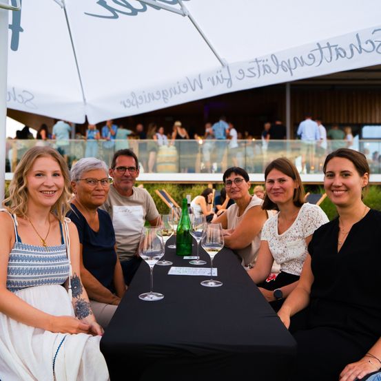 Fünf Frauen und ein Mann sitzen um einen Tisch und lächeln für ein Foto. Auf dem Tisch stehen Weingläser, eine Flasche und eine Karte. Hinter ihnen sitzen und stehen Leute auf einem Balkon. Ein großer Regenschirm ist über ihnen.