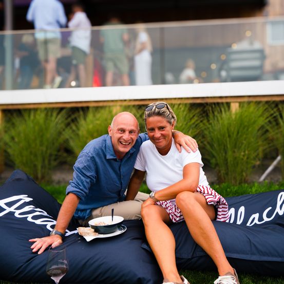 Ein Mann und eine Frau sitzen auf einem Beanbag-Sessel auf einer Rasenfläche. Sie lächeln und schauen in die Kamera. Hinter ihnen zeigt ein unscharfer Hintergrund eine Glaswand mit Menschen und einem Gebäude.
