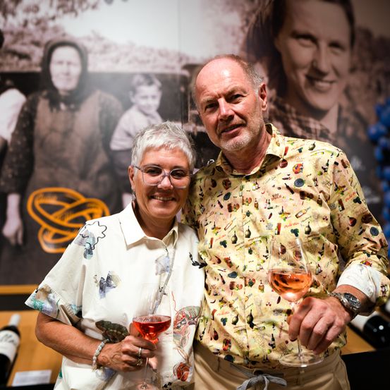 Ein Mann und eine Frau lächeln und halten Weingläser in der Hand, stehen vor einem Fotohintergrund mit einer älteren Frau, einem Kind und einem lächelnden Mann.