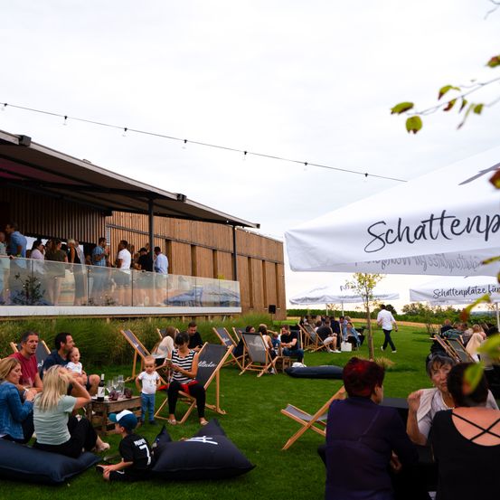 Ein Außenbereich mit Menschen, die auf Beanbags und Stühlen sitzen. Ein Gebäude mit Balkon im Hintergrund. Ein weißer Schirm mit dem Text Schattenplatz rechts.