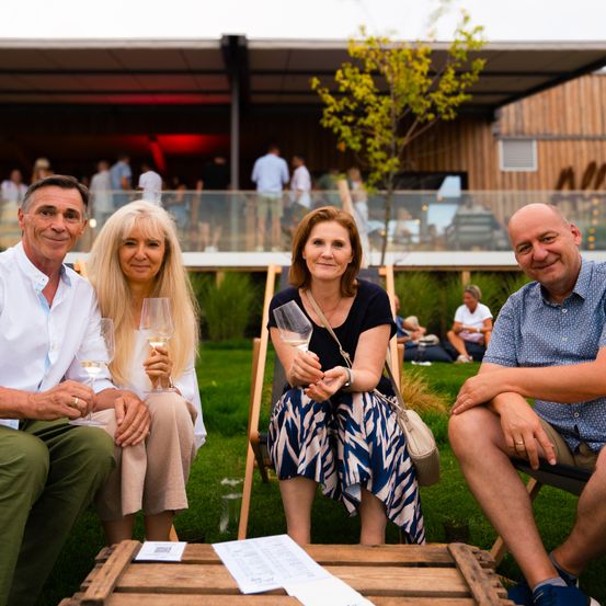 Vier Personen, zwei Männer und zwei Frauen, sitzen auf Stühlen im Freien. Sie halten Weingläser und lächeln. Sie sind von einer Rasenfläche und einer Holzstruktur mit einer Glaswand umgeben.