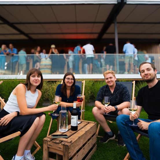 Vier Personen sitzen auf Stühlen um einen Holzkasten herum, lächeln und halten Weingläser. Hinter ihnen steht eine Gruppe von Menschen an einem Glasgeländer und beobachtet. Der Ort ist draußen, mit einem Rasenbereich und einem modernen Gebäude im Hintergrund.