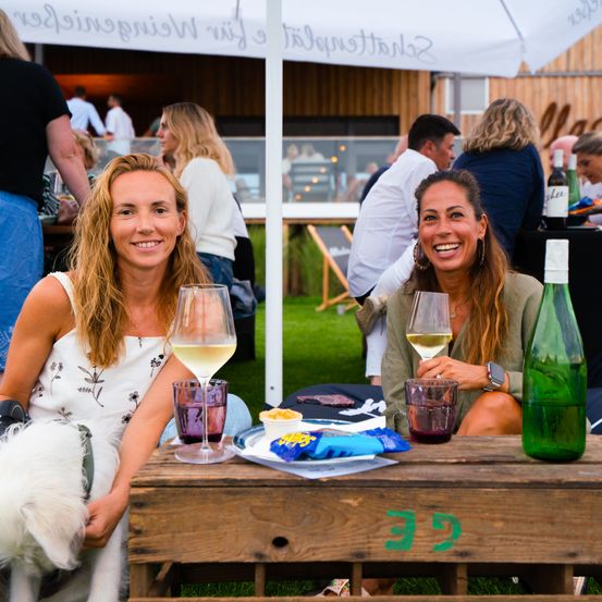 Zwei Frauen mit Weingläsern sitzen auf einem Holztisch im Freien mit einem Hund. Hinter ihnen sitzen und stehen Leute unter einem Zelt. Eine Frau hält eine Flasche Wein, während die andere eine Flasche Sekt hat.