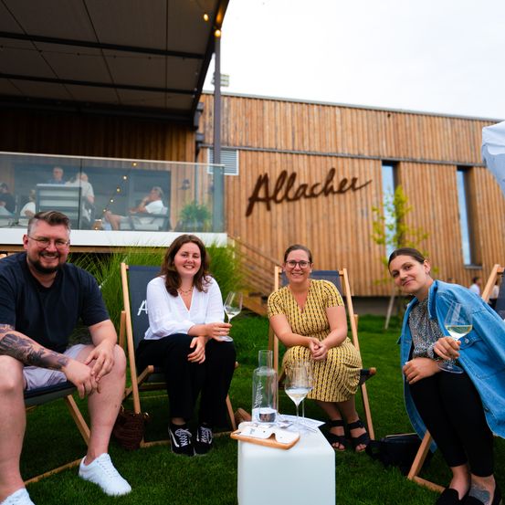 Vier Personen sitzen auf Stühlen vor einem Gebäude mit einem Schild, das Allacher steht. Zwei Frauen und ein Mann lächeln und halten Weingläser. Eine weitere Frau hat ein Getränk auf einem Tisch.