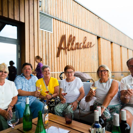 Eine Gruppe von Erwachsenen sitzt auf einer Couch, hält Weingläser in der Hand und lächelt. Die hölzerne Wand hinter ihnen hat den Schriftzug 'Allacher' darauf. Auf dem Tisch stehen Weinflaschen, und ein Kinderwagen ist in der Nähe geparkt. Eine Person steht hinter der Gruppe.