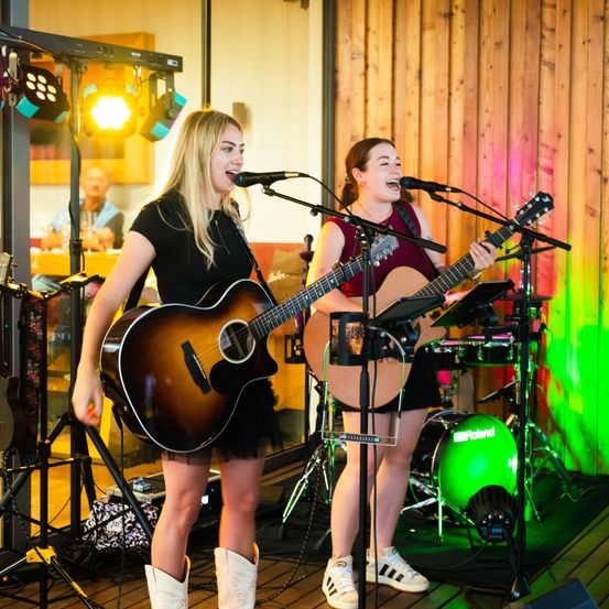 Zwei Frauen spielen Gitarre und singen auf der Bühne. Sie sind von Bühnenlichtern beleuchtet, mit einem Schlagzeug und Becken dahinter. Die Bühne hat Holzwände und einen Holzboden. Ein Mann sitzt am Tisch im Hintergrund.