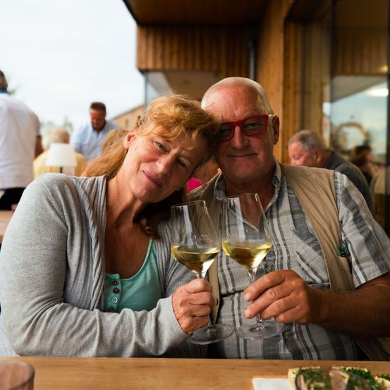 Ein älteres Paar mit Weinglas sitzt auf einer Bank. Der Mann trägt eine Brille und eine Weste. Hinter ihnen sitzen weitere Personen auf Bänken. Links steht eine Lampe auf einem Tisch.