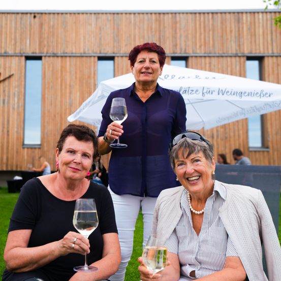 Drei Frauen in einem Garten, eine hält ein Weinglas, eine andere hält zwei, und eine dritte lächelt und steht hinter ihnen.