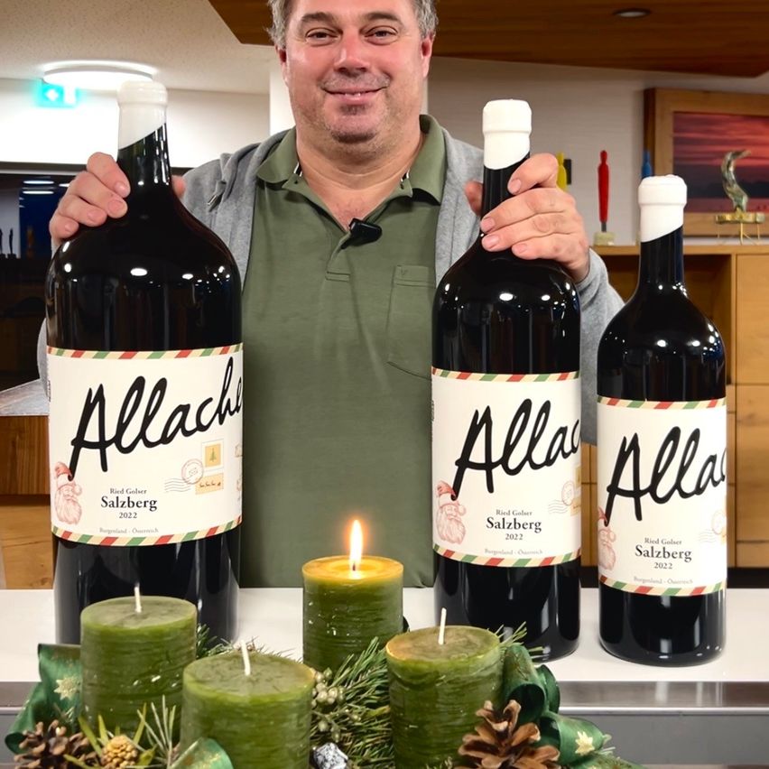 Ein Mann hält drei Flaschen Wein mit dem Aufkleber Allach Salzburg 2022 auf einem Tisch mit Kerzen und Tannenzapfen.