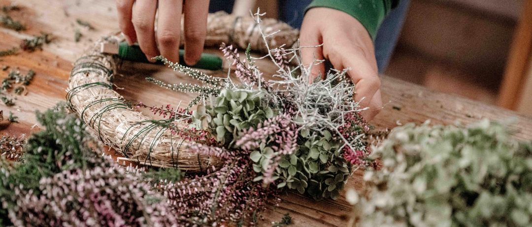 Eine Person in einem grünen Langarmshirt stellt einen Kranz aus getrockneten Blumen und Grünzeug auf einem Holztisch her. Der Kranz hat eine kreisförmige Form und eine grüne Schleife.
