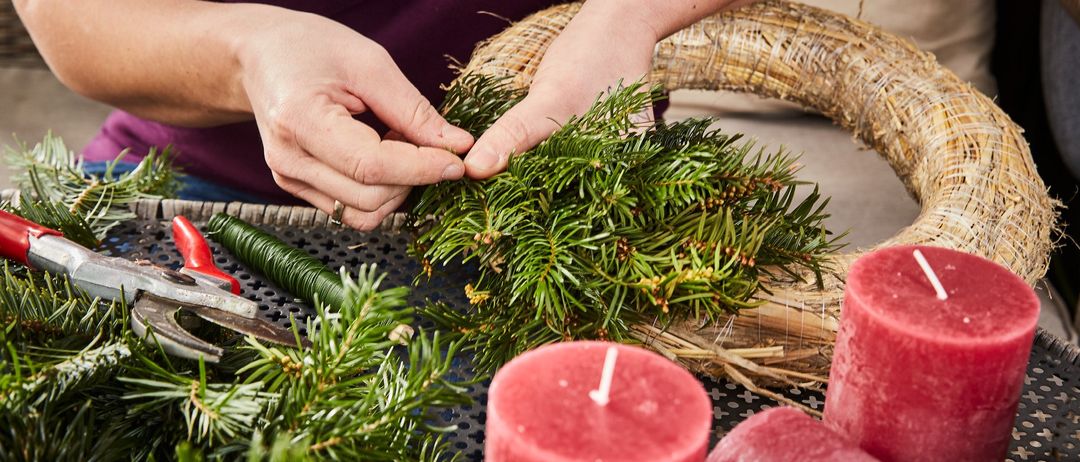 Eine Person dekoriert einen Kranz mit Tannenzweigen, benutzt eine Schere und drei rote Kerzen. Die Szene spielt sich auf einer blauen Oberfläche ab.