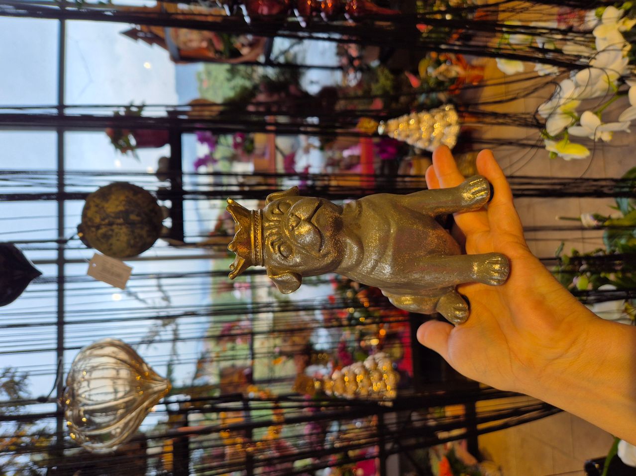 Eine Person hält eine bronzene Bulldoggenfigur mit Krone, vor dem Hintergrund eines Geschäfts mit verschiedenen Dekorationsartikeln.