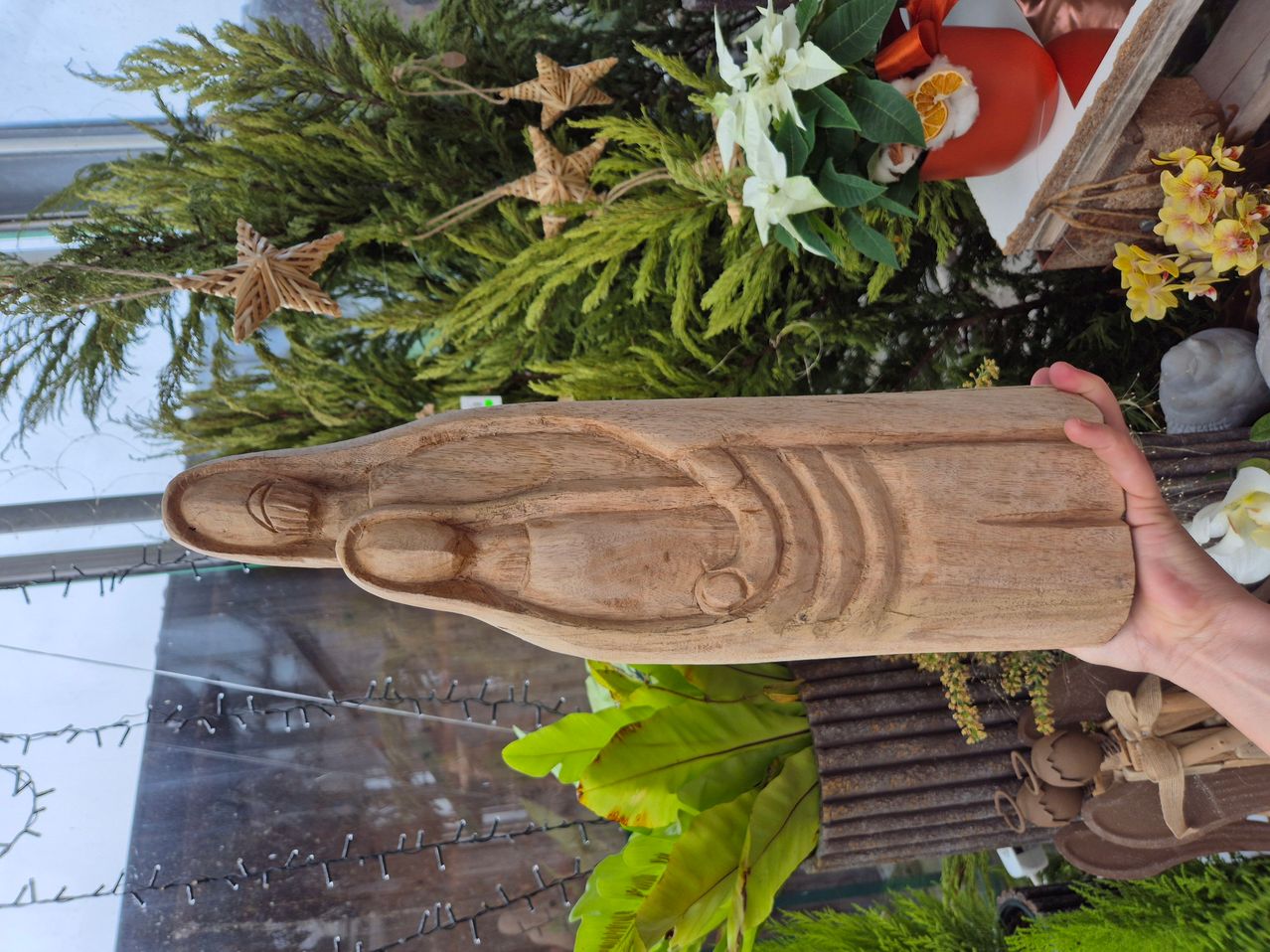 Eine Holzskulptur einer Frau wird vor einem Hintergrund aus Weihnachtsdekoration und Pflanzen hochgehalten.