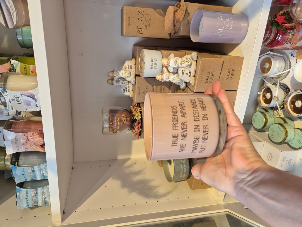 Eine Hand hält eine Kerze mit der Aufschrift 'Wahre Freunde sind nie getrennt, vielleicht in der Ferne, aber nie im Herzen'. Dahinter stehen verschiedene Gegenstände auf einem Regal, darunter eine Schachtel 'Relax' Kerzen.