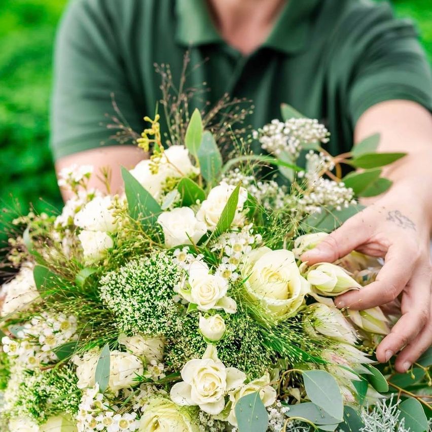 Eine Person in einem grünen Poloshirt hält einen großen Strauß weißer Blumen und Grün. Der Strauß besteht aus weißen Rosen, grünen Blättern und kleinen weißen Blüten. Der Hintergrund scheint draußen zu sein, mit einem grünen Hintergrund.