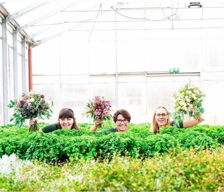 Drei Frauen posieren in einem Gewächshaus, jede hält einen Blumenstrauß, mit sichtbarem Metallrahmen und klarem Dach.