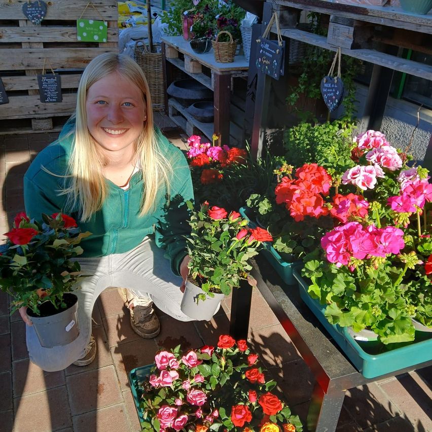 Eine lächelnde Frau in Grün hält eine Topfpflanze in einem Blumenladen, umgeben von verschiedenen Blumen, Regalen und Lagerboxen.