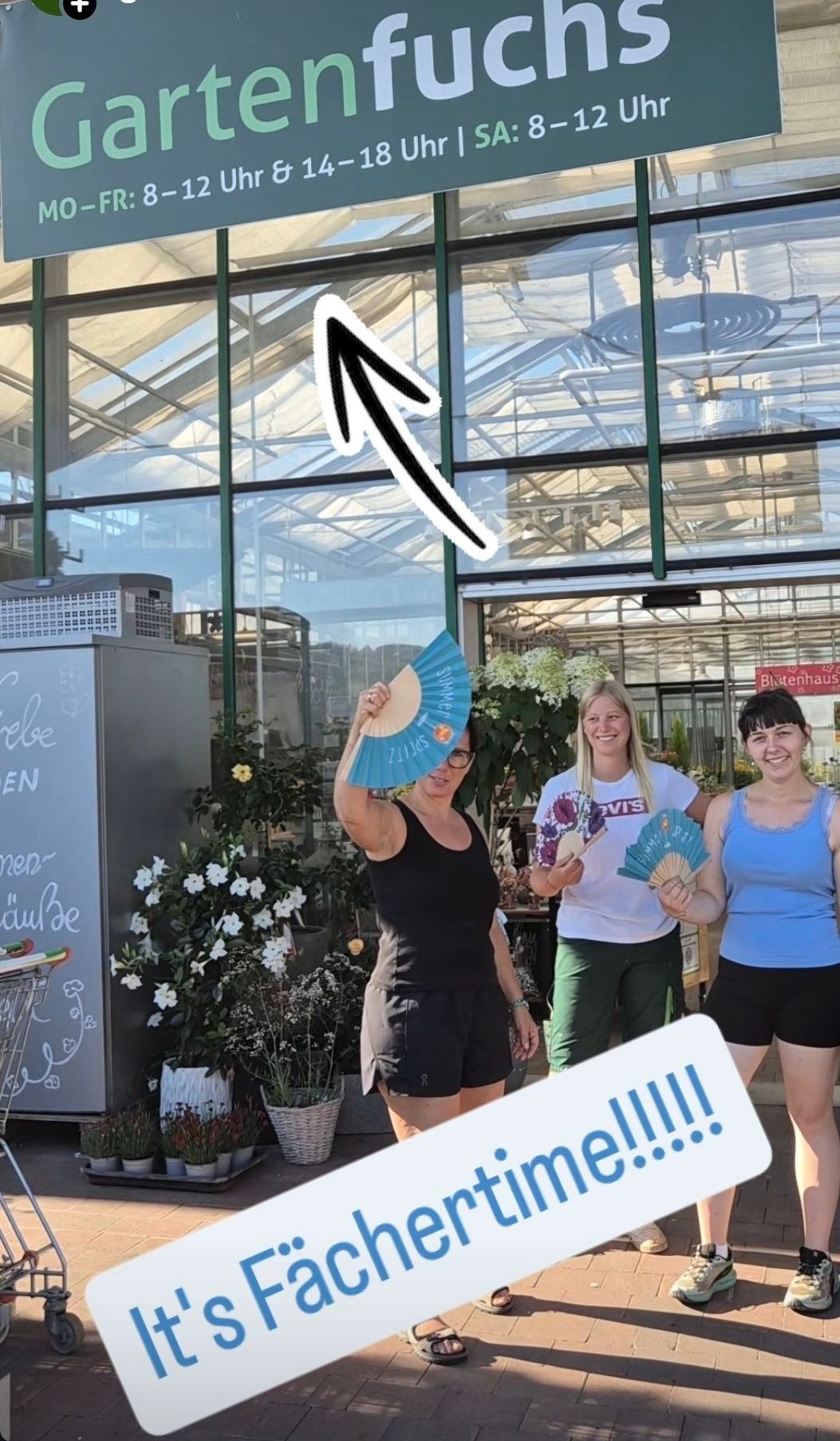 Drei Frauen stehen vor einem Gewächshaus, eine hält einen Fächer. Die mittlere Frau trägt ein weißes T-Shirt, die anderen Schwarz und Blau. Sie stehen neben einem Einkaufswagen und Blumen.