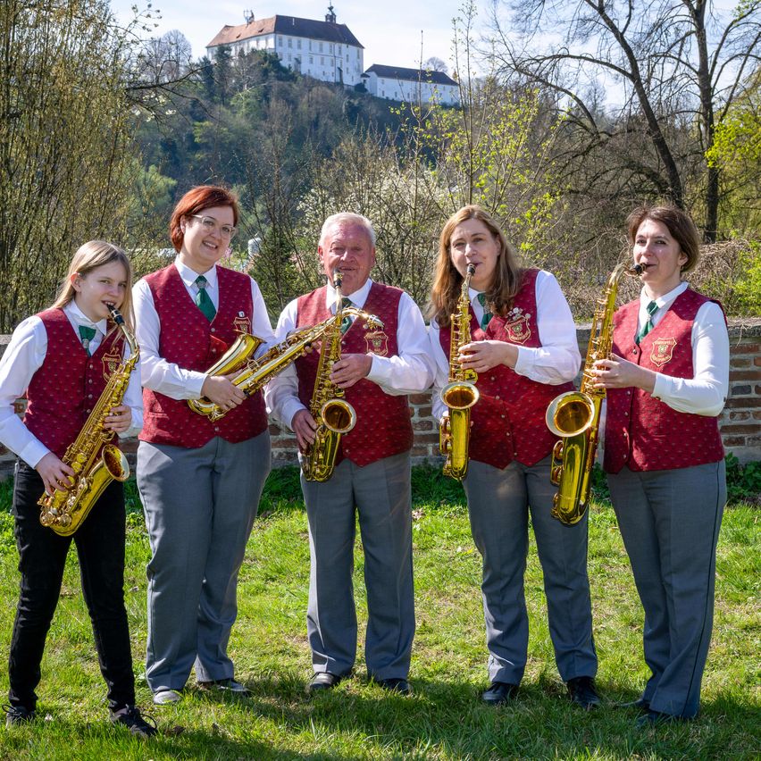 Fünf Menschen stehen auf dem Rasen und posieren für ein Foto. Sie halten alle Saxophone. Die Frau links trägt eine Brille. Das Schloss befindet sich auf dem Hügel. Der Himmel ist klar.